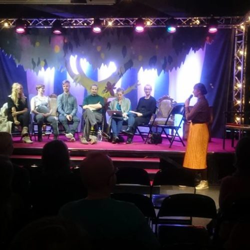 Pienellä näyttämöllä istuu kuusi panelistia rinnakkain. Juontaja Veera Lamberg seisoo ja puhuu mikrofoniin.