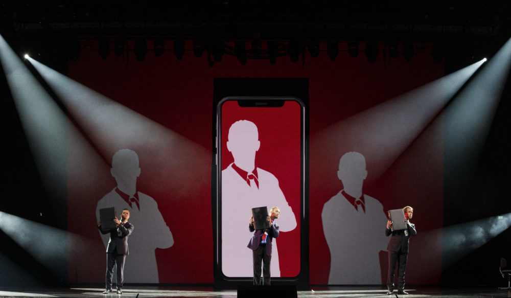 Näyttämöllä on valtava kännykkä, jonka edessä kolme näyttelijää elehtii läppärit käsissään.