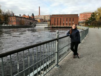 Anu Järvensivu nojailee metallikaiteeseen Tammerkosken rannalla, hiekkatiellä. Taustalla näkyy Tammerkoski ja vanhat punatiilirakennukset kosken molemmin puolin.