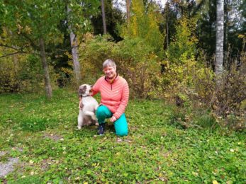 Sikke Leinikki kyyköttää pihamaalla koiransa Maran kanssa. Leinikillä on punaiset silmälasinsangat, punainen tikkitakki ja turkoosinsiniset housut.