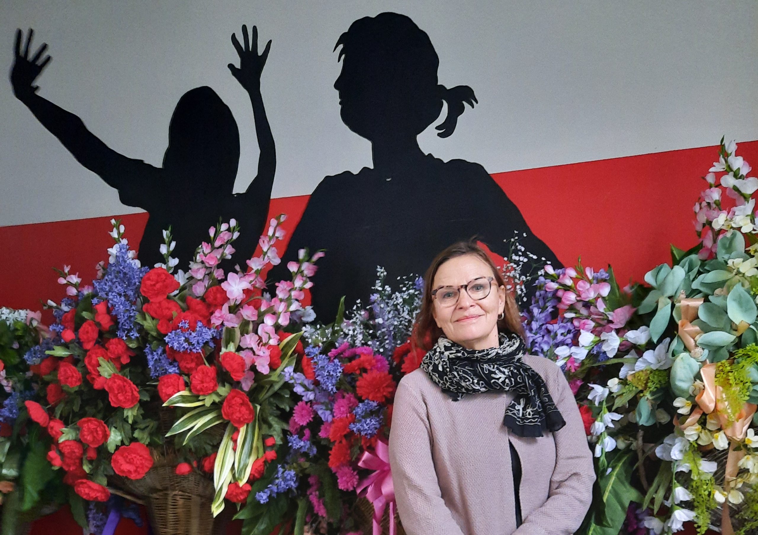 Museoteatterin johtaja hymyilee kameralle Teatterimuseon sosiaalitilassa, jonka seinälle on piirretty kahden henkilön siluetit. Seilon taustalla, seinän vieressä on paljon tekokukkia.