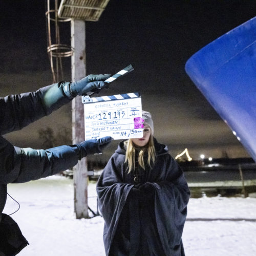 Venesatamassa, lumen keskellä elokuvatyöntekijä näyttää klaffia vaaleatukkaisen näyttelijättären edessä. Oikealla näkyy sinistä venettä.