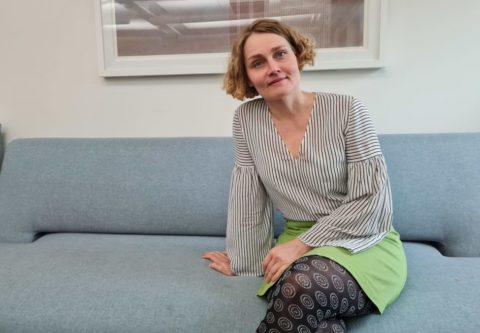 Susanna Metsälä istuu sinisellä sohvalla mustavalkoraidallisessa paidassa, vihreässä hameessa ja kuviollisissa tummanruskeissa sukkahousuissa.